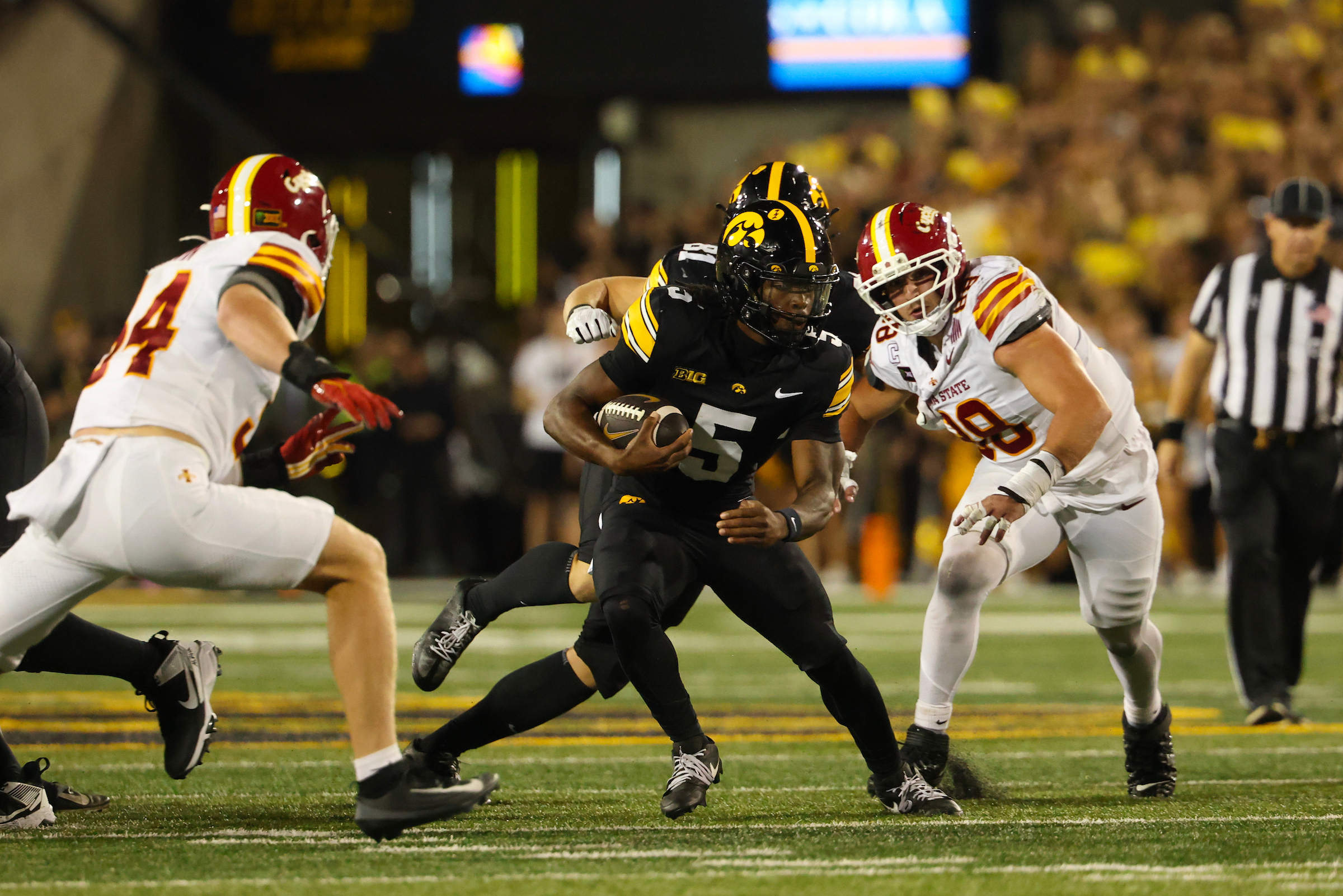 A football player in a black uniform with the number 5 runs with the ball, being pursued by two opponents in white uniforms during a game.