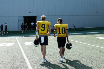 Two athletes in yellow jerseys walk away from the camera across a football field, each carrying a helmet, with a large building in the background.