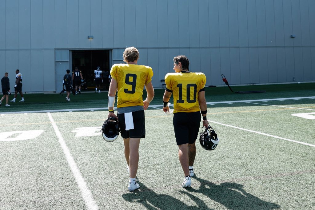 Two athletes in yellow jerseys walk away from the camera across a football field, each carrying a helmet, with a large