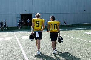 Two athletes in yellow jerseys walk away from the camera across a football field, each carrying a helmet, with a large building in the background.