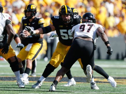 A football player in a black and yellow uniform with the number 55 is engaged in a play, facing off against an opponent wearing number 97 on a white jersey.
