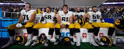 Five American football players in white and yellow uniforms sit on a bench during a game, with helmets and gear nearby.