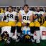 Five American football players in white and yellow uniforms sit on a bench during a game, with helmets and gear nearby.