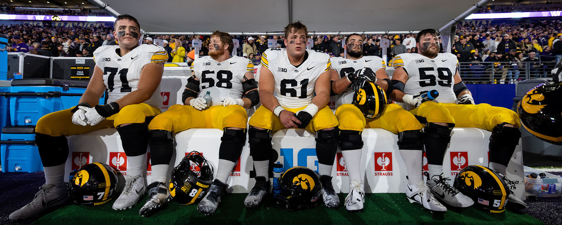 Five American football players in white and yellow uniforms sit on a bench during a game, with helmets and gear nearby.
