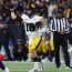 A football quarterback in a white and yellow uniform throws the ball while being defended by players in purple uniforms on a field with a large crowd in the background.
