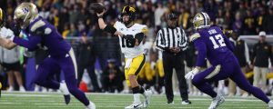 A football quarterback in a white and yellow uniform throws the ball while being defended by players in purple uniforms on a field with a large crowd in the background.